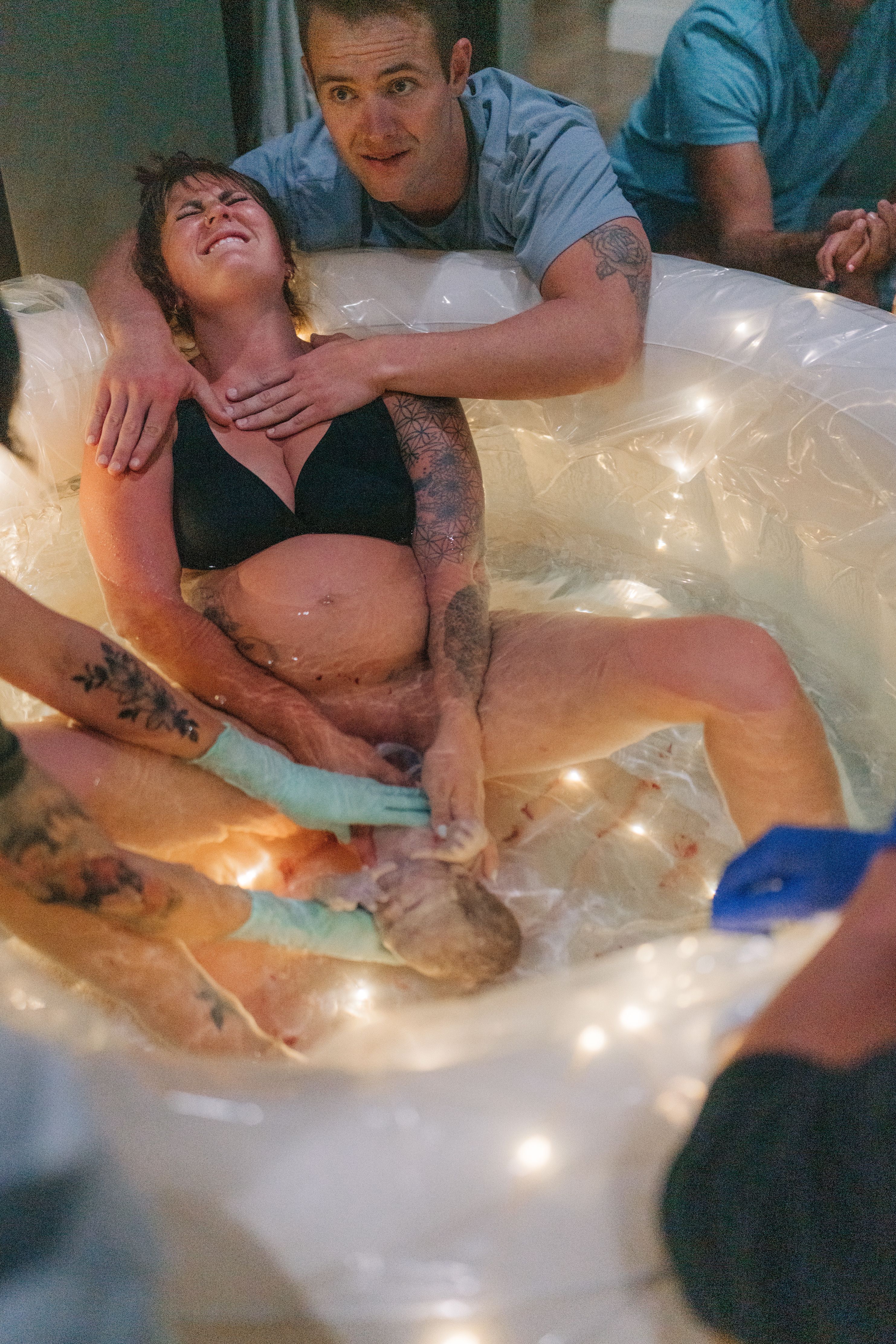 A mother giving birth in a softly lit birth pool, her partner holding her from behind
