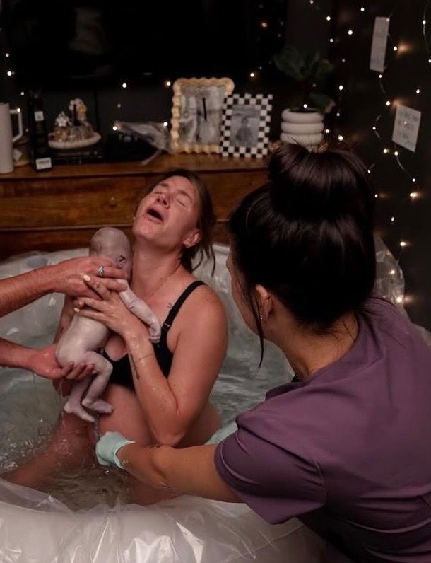 A mother in a birth pool lifting her newborn to her chest as a midwife supports her