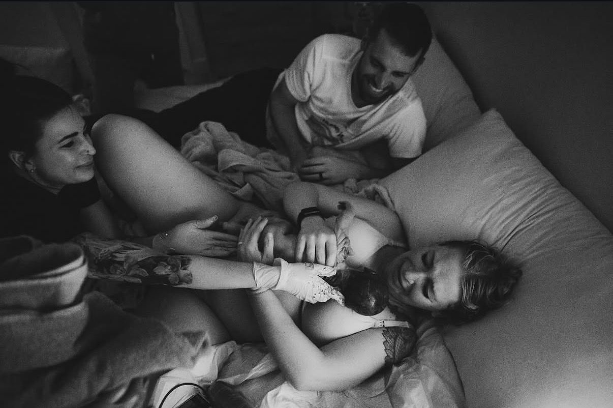 A mother resting in bed with her newborn on her chest as her partner and midwife smile beside her